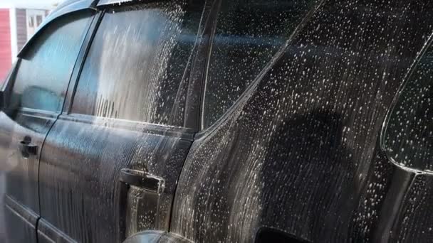 Autowaschvorgang an einer autowaschanlage zur selbstbedienung. Ein wasserstrahl mit hohem druck wäscht den schaum vom auto ab. Seitenansicht. geschäumtes Waschmittel fließt von der Karosserieoberfläche ab — Stockvideo