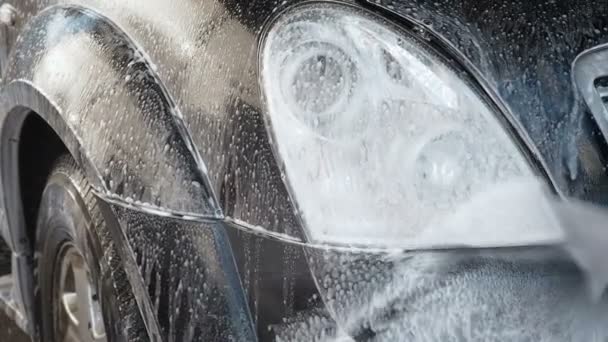 Video en cámara lenta de un proceso de lavado de autos en un auto-servicio de lavado de autos. Un chorro de agua con un lavado de alta presión de la suciedad del faro del auto. Vista lateral. El detergente espumado drena de la superficie — Vídeo de stock