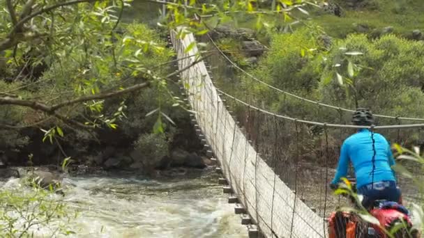 Cycliste monte un pont suspendu étiré à travers une rivière de montagne orageuse. Un cycliste roule sur un vélo le long d'un long pont suspendu étroit en bois qui s'étend entre les rives d'un turbulent — Video