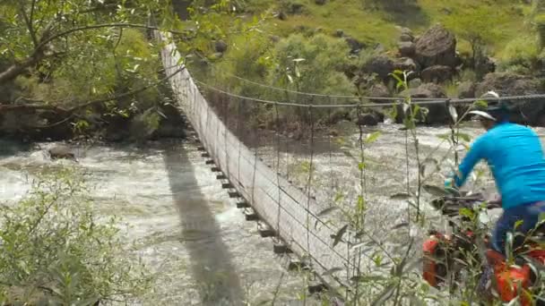 Bicyclist rides a suspension bridge stretched across a stormy mountain river. A cyclist rides on a bicycle along a long narrow wooden suspension bridge that stretches between the banks of a turbulent — Stock Video