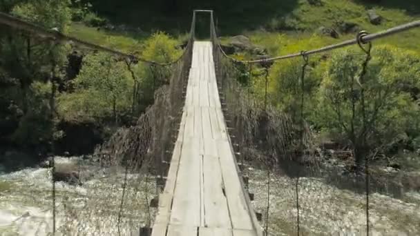 Atravessando para o outro lado de um rio de montanha por uma ponte de madeira suspensa. Ponte de suspensão passeio de rio de montanha — Vídeo de Stock