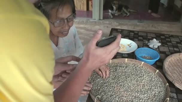 Provincia de Chiang Mai, Tailandia - 23 de mayo de 2019: Un hombre muestra algo a la anciana en la pantalla de un teléfono inteligente, sonríe en proceso de clasificación a través de granos de café árabe en mimbre redondo pequeño — Vídeo de stock