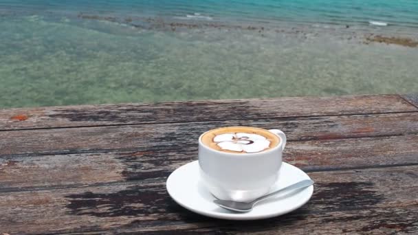 Geflavoreerde koffie in witte kop op plaat met lepel op houten rustieke tafel, zee achtergrond met de zee golven beweging. Koffie drinken met uitzicht op zee in een tropisch resort. Morning Cup van geflavoreerde — Stockvideo