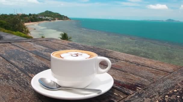 Café na taça branca na placa com colher na mesa rústica de madeira, fundo do mar com o movimento da palmeira. Fundo de verão bonito — Vídeo de Stock
