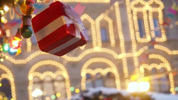 Anno nuovo e scenario stradale di Natale della città europea. La confezione regalo decorativa legata con un nastro bianco satinato ruota su una corda sospesa all'abete rosso sullo sfondo luminoso illuminato — Video Stock