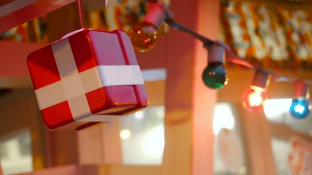 Año Nuevo y Navidad paisaje callejero de la ciudad europea. La caja de regalo decorativa atada con una cinta blanca satinada gira sobre una cuerda suspendida de abeto sobre el fondo iluminado brillante — Vídeos de Stock