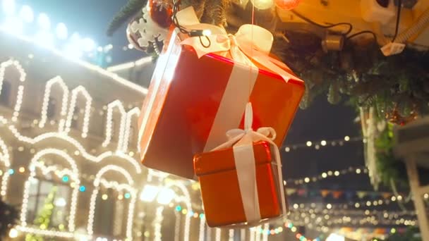 Año Nuevo y Navidad paisaje callejero de la ciudad europea. La caja de regalo decorativa atada con una cinta blanca satinada gira sobre una cuerda suspendida de abeto sobre el fondo iluminado brillante — Vídeo de stock