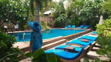 Mavi mayo, şapka ve güneş gözlüğü takmış güzel bir bayan güneşli bir günde temiz suyla yüzme havuzunda dolaşıyor. Lüks tatil köyü yeşillik içinde boğuluyor. Mavi güneşli gölgelerde yüzme anketi