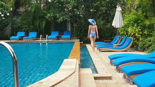 Bella dama en traje de baño azul, sombrero y gafas de sol caminando alrededor de la piscina con agua clara en un día soleado. Resort de lujo ahogándose en la vegetación. Encuesta de natación con tumbonas azules a la sombra de — Vídeo de stock