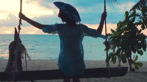 Beautiful girl in a hat at sunset swinging on a swing on a seashore. Silhouette pretty young woman sitting on a hanging swing and admiring sea sunset on a tropical island — Stock Video