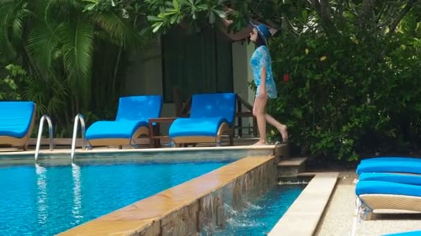 Hübsche Dame in blauem Badeanzug, Hut und Sonnenbrille spaziert und liegt an einem sonnigen Tag auf einer Liege am Pool mit klarem Wasser. Luxusresort ertrinkt im Grünen. Schwimmen mit Blau — Stockvideo