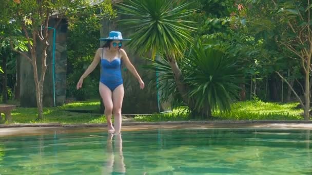 Chica sllim sexy en un traje de baño azul, sombrero y gafas de sol entra en la piscina termal en los escalones. Lento movimiento Jovencita entra en la piscina verde esmeralda con agua caliente. Mujer pone su pie en el claro — Vídeos de Stock