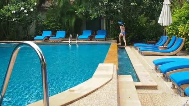 Bella dama en traje de baño azul, sombrero y gafas de sol caminando alrededor de la piscina con agua clara en un día soleado. Resort de lujo ahogándose en la vegetación. Encuesta de natación con tumbonas azules a la sombra de — Vídeo de stock