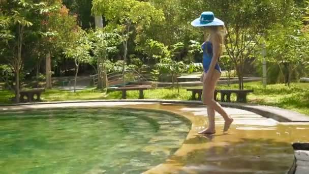 Sexy ragazza sllim in costume da bagno blu, cappello e occhiali da sole entra nella piscina termale sui gradini. Rallentatore Giovane signora entra nella piscina verde smeraldo con acqua calda. Donna mette il piede nel limpido — Video Stock