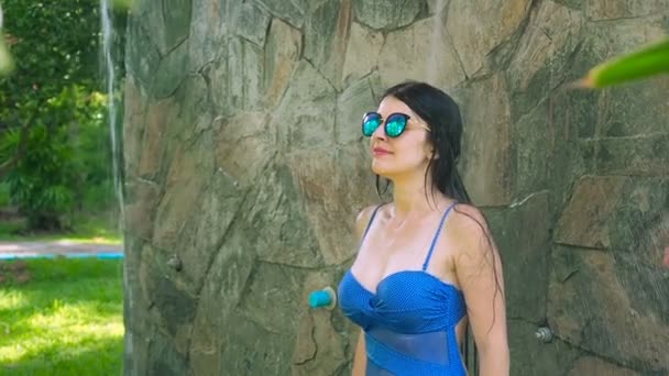 Sexy brunette woman in blue one-piece swimsuit and sunglasses taking outdoor shower at the resort in the tropics slowmotion — Stock Video