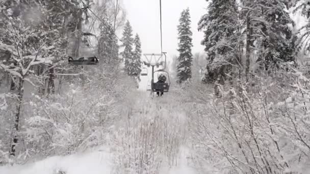 Aerial view of a two-seat cable car over the winter snowy forest. Chairlift climb above the ski slope in small ski resort Stepanovo during a snowfall — ストック動画