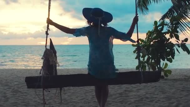 Vacker flicka i hatt vid solnedgången svingande på en gunga på stranden. Silhuett söt ung kvinna sitter på en hängande gunga och beundrar havet solnedgång på en tropisk ö — Stockvideo