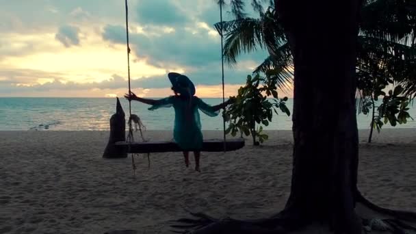 아름다운 아가씨가 석양에 모자를 쓴 채 해변에서 그네를 타고 있습니다. 아주 젊은 여자가 매달려 그네를 타고 열 대의 섬에서 해가 지는 것을 감상하며 앉아 있는 모습 — 비디오
