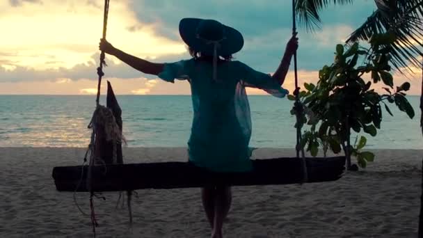 Gyönyörű lány kalapban naplementekor hintán hintázva a tengerparton. Silhouette csinos fiatal nő ül egy lógó hinta és csodálta a tengeri naplemente egy trópusi szigeten — Stock videók