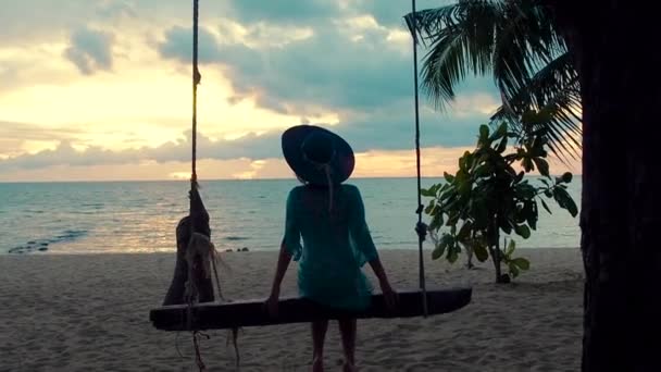 Красивая девушка в шляпе на закате качается на качелях на берегу моря. Силуэт красивая молодая женщина сидит на висячих качелях и любуется закатом моря на тропическом острове — стоковое видео