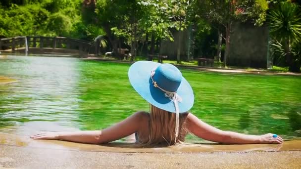 Bella dama en un traje de baño azul, sombrero y gafas de sol se sienta y se relaja en la piscina tropical con agua clara en una vista posterior día soleado. Mujer endereza el sombrero en el agua verde de la piscina tropical — Vídeo de stock