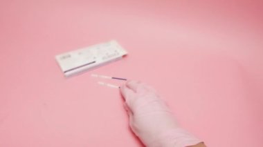 Pozitif hamilelik testi tutan pembe tıbbi eldivenli bir kadın eli ve pembe arka planda izole edilmiş bir jest gösteriyor. Mavi çubuktaki HCG kısaltması insan koryonik gonadotropininin...