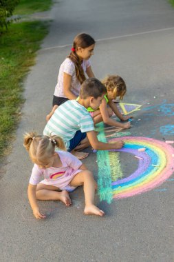 Çocuklar asfalta tebeşirle resim çizerler. Gökkuşağı, ev, güneş, bulutlar asfalta çizilmiş..