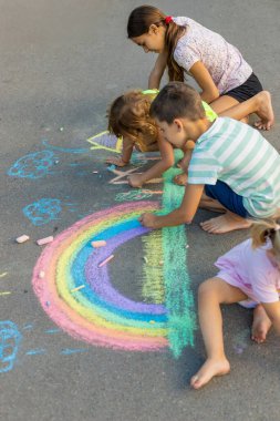 Çocuklar asfalta tebeşirle resim çizerler. Gökkuşağı, ev, güneş, bulutlar asfalta çizilmiş..