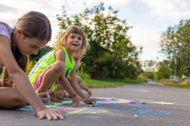 Çocuklar asfalta tebeşirle resim çizerler. Gökkuşağı, ev, güneş, bulutlar asfalta çizilmiş..