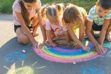 Çocuklar asfalta tebeşirle resim çizerler. Gökkuşağı, ev, güneş, bulutlar asfalta çizilmiş..