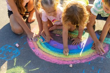 Çocuklar asfalta tebeşirle resim çizerler. Gökkuşağı, ev, güneş, bulutlar asfalta çizilmiş..