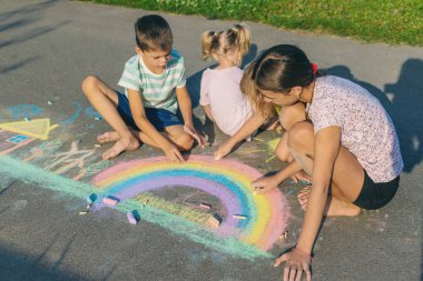 Çocuklar asfalta tebeşirle resim çizerler. Gökkuşağı, ev, güneş, bulutlar asfalta çizilmiş..