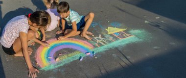 Çocuklar asfalta tebeşirle resim çizerler. Gökkuşağı, ev, güneş, bulutlar asfalta çizilmiş..