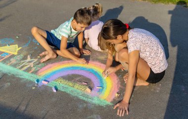 Çocuklar asfalta tebeşirle resim çizerler. Gökkuşağı, ev, güneş, bulutlar asfalta çizilmiş..