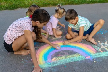 Çocuklar asfalta tebeşirle resim çizerler. Gökkuşağı, ev, güneş, bulutlar asfalta çizilmiş..