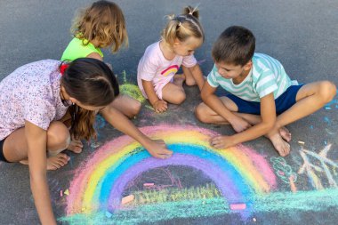 Çocuklar asfalta tebeşirle resim çizerler. Gökkuşağı, ev, güneş, bulutlar asfalta çizilmiş..