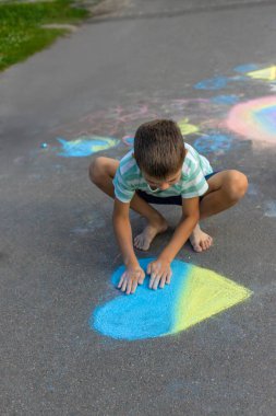 Bir çocuk tebeşirle bir kalp çiziyor ve onu maviye ve sarıya boyuyor..
