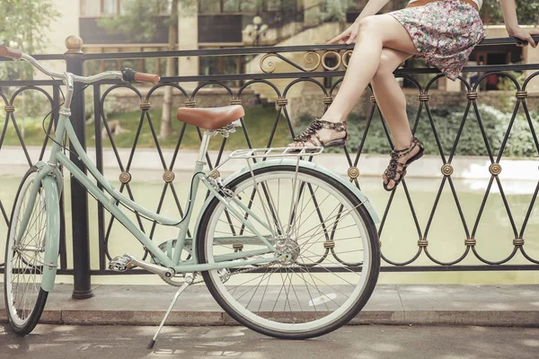 Молодая девушка сидит на заборе возле винтажного велосипеда в парке — стоковое фото