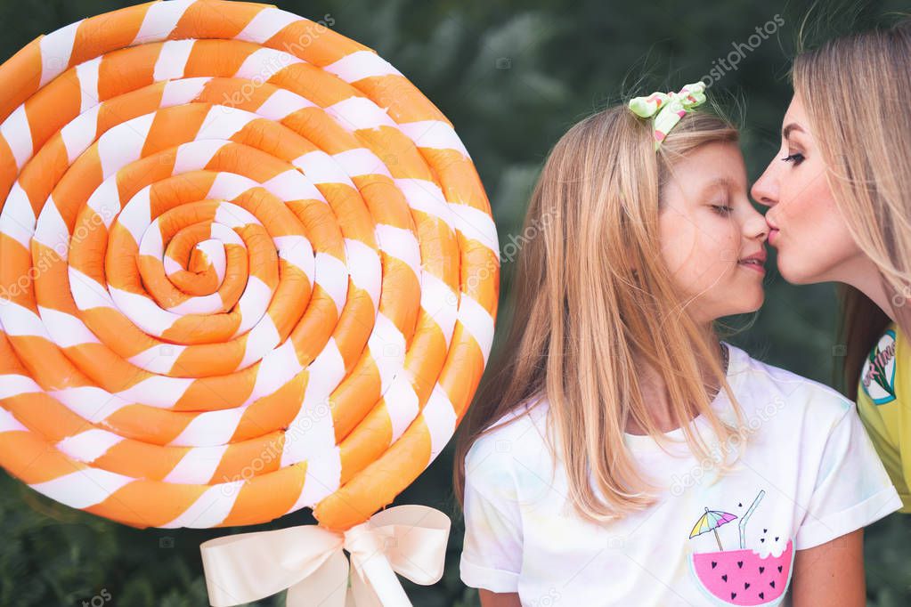 Blonde Mum Kissing Her Daughter. Girl Holding Big Chupa Chups