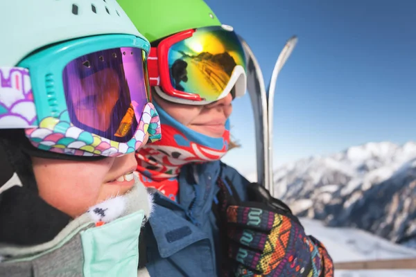 Feliz mamá y niña sentadas en la silla en el telesilla sonriendo y usando máscaras de esquí con la montaña en el fondo —  Fotos de Stock