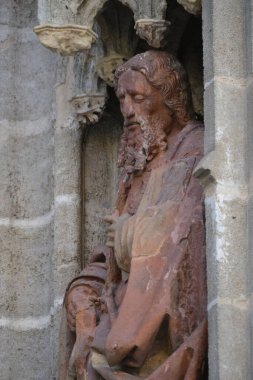 A detailed stone sculpture of a man with a beard and long hair, weathered and reddish in color, is set within a Gothic architectural niche. The figure looks downward with a somber expression.