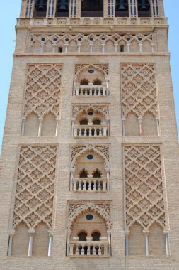 A vertical shot of a tall brick tower shows intricate Moorish-style architecture. The facade is detailed with carved patterns, arched windows, and small balconies, rising towards the top where bells are visible.