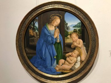 This is a Renaissance tondo painting, depicting the Madonna and Child. The Virgin Mary, dressed in a blue robe, is shown with hands together in prayer, gazing down at a reclining baby Jesus. Another child figure is also present, with all three figure