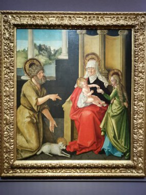 A painting in a gilded frame shows a religious scene. A man in a brown robe kneels on the left, gesturing toward a woman in a red dress who holds a baby on her lap. To the right of the woman stands a young girl, and a small lamb is on the floor.