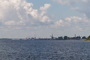 Ufukta kargo vinçleri ve fabrika binaları olan sanayi limanı manzarası bulutlu bir gökyüzünün altında suyun üzerinde görülüyor..
