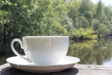 Güneşli bir yaz gününde tahta bir masada beyaz bir kupa. Günün başlangıcına ait huzurlu bir resim. Güzel bir günün sabahı.