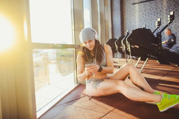 Deportes temáticos, salud y tecnología. hermosa mujer sexy caucásica deportista en ropa deportiva gris y sombrero se sienta por la ventana con puesta de sol utiliza teléfono inteligente escuchar música en auriculares grandes en la cabeza — Foto de Stock