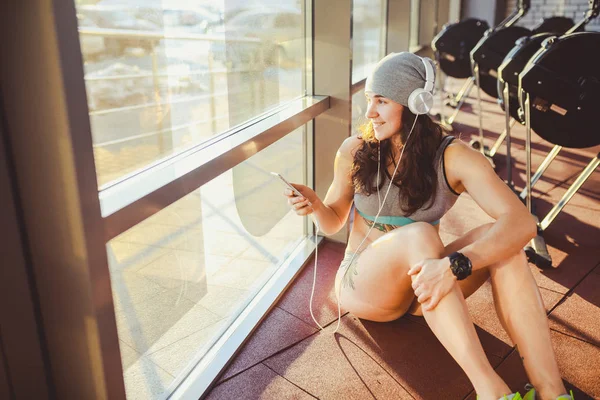 主题体育, 健康和技术。美丽性感的白种女人运动员在灰色运动服和帽子坐在窗口与太阳设置使用智能手机听音乐在大耳机头上 — 图库照片