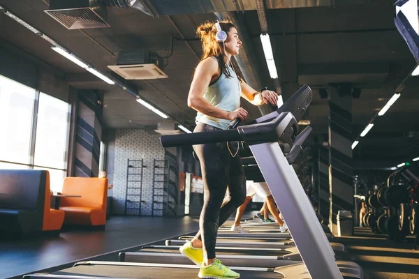 Beautiful young woman with long hair trains in the gym on a treadmill. Listens to music in large headphones. On his arm a sports watch in black for tracking speed, heart rate, pace and stopwatch — Stock Photo, Image