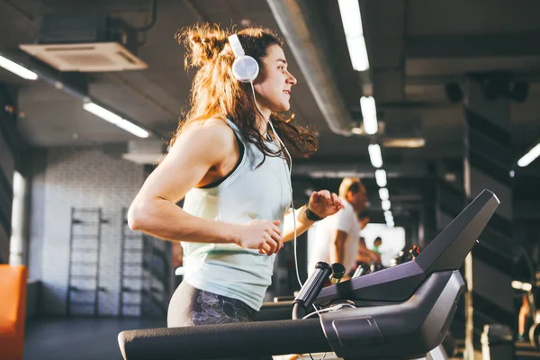 El tema es deporte y música. Una hermosa mujer inflada corre en el gimnasio en una cinta de correr. En su cabeza hay grandes auriculares blancos, la chica escucha música durante un entrenamiento cardiovascular para bajar de peso — Foto de Stock